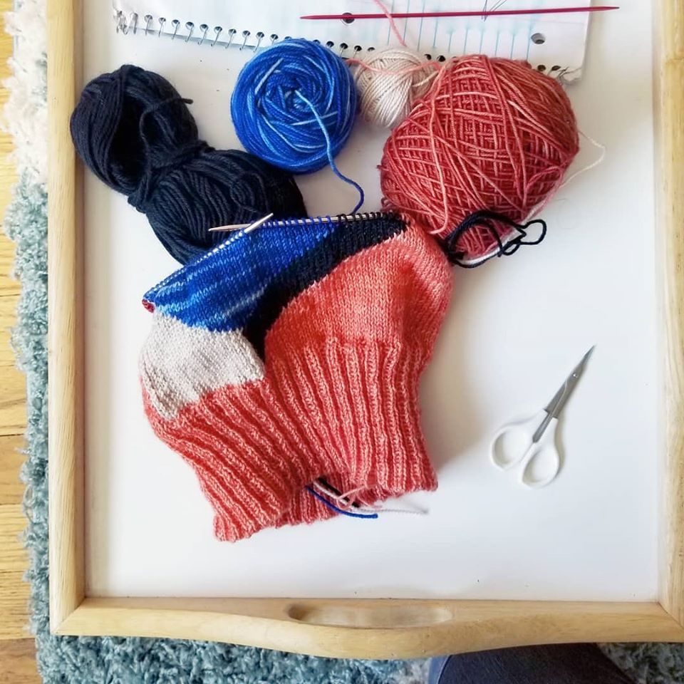 A knitting project in progress, laid out on a table. The colors are pink, off-white, dark blue, and bright blue arranged in a geometric colorblock pattern.