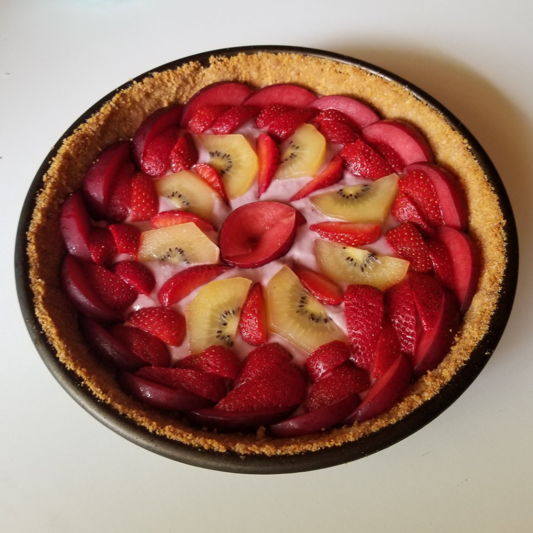 Photo of a fruit tart in a pie tin, topped with slices of fresh plums, kiwis, and strawberries.